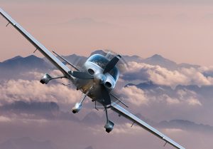 front view of a single prop airplane flying against clouds and mountains
