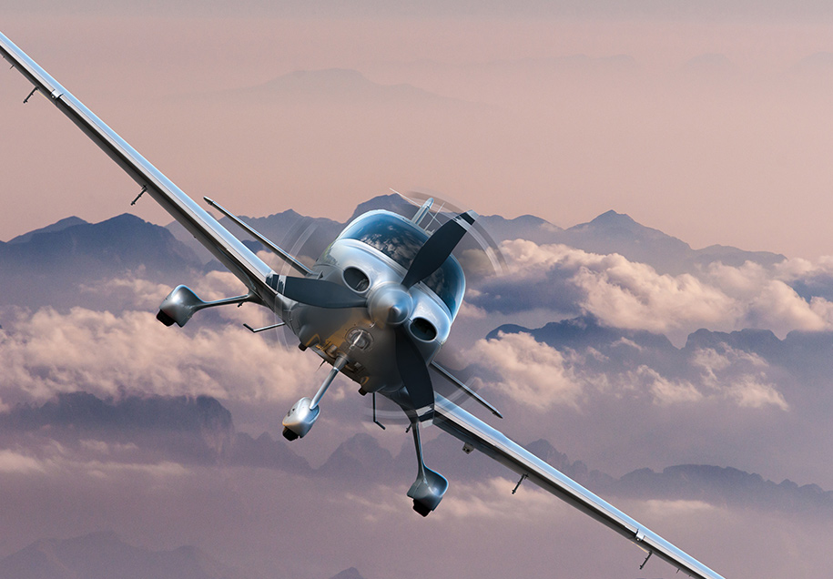 front view of a single prop airplane flying against clouds and mountains